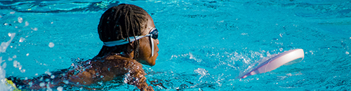A girl swimming in a pool holding a small kick board and wearing swimming goggles.